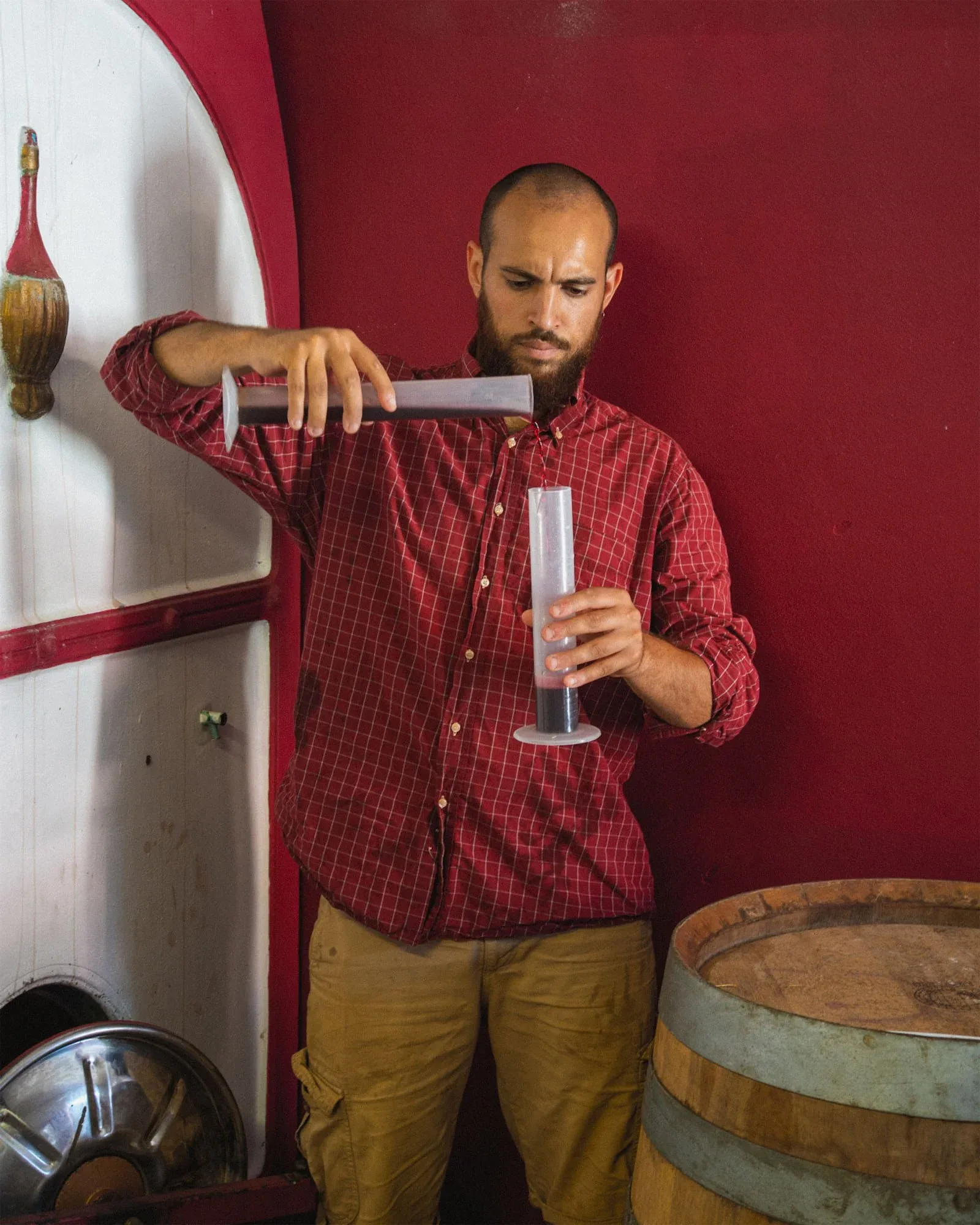 Uomo in camicia a quadri che versa il vino in un cilindro graduato accanto a una botte, davanti a un muro rosso: foto dallo shooting fotografico per Colle Vento