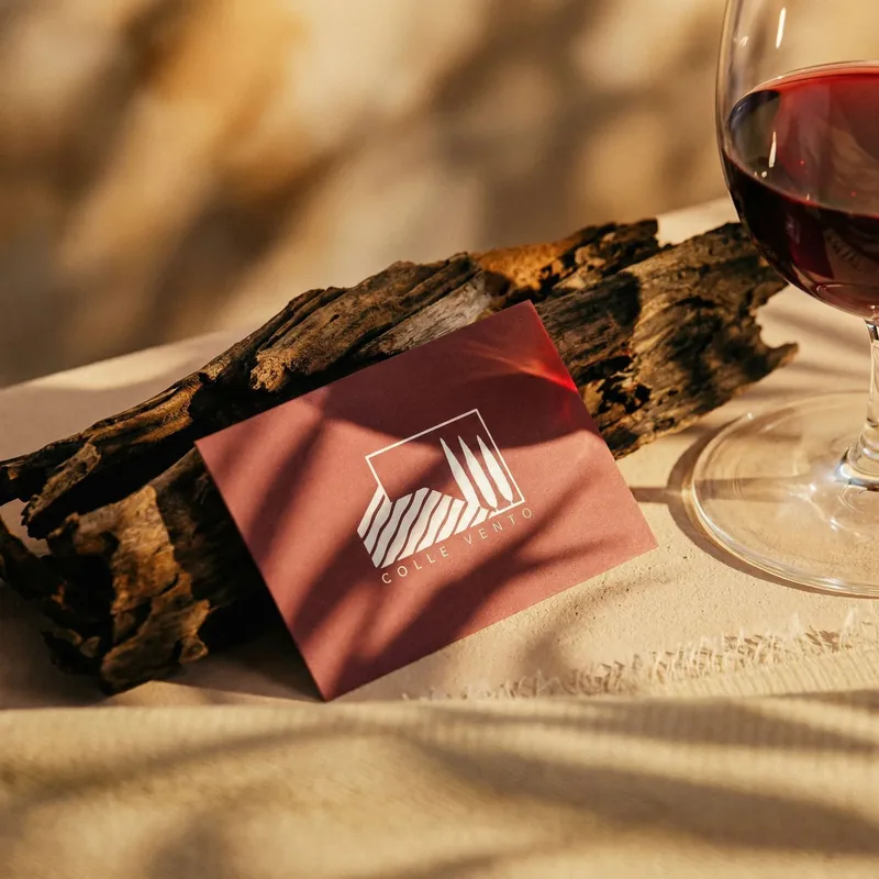 Biglietto da visita bordeaux col logo Colle Vento appoggiato a un pezzo di corteccia, accanto a un calice di vino rosso: mockup di brand identity