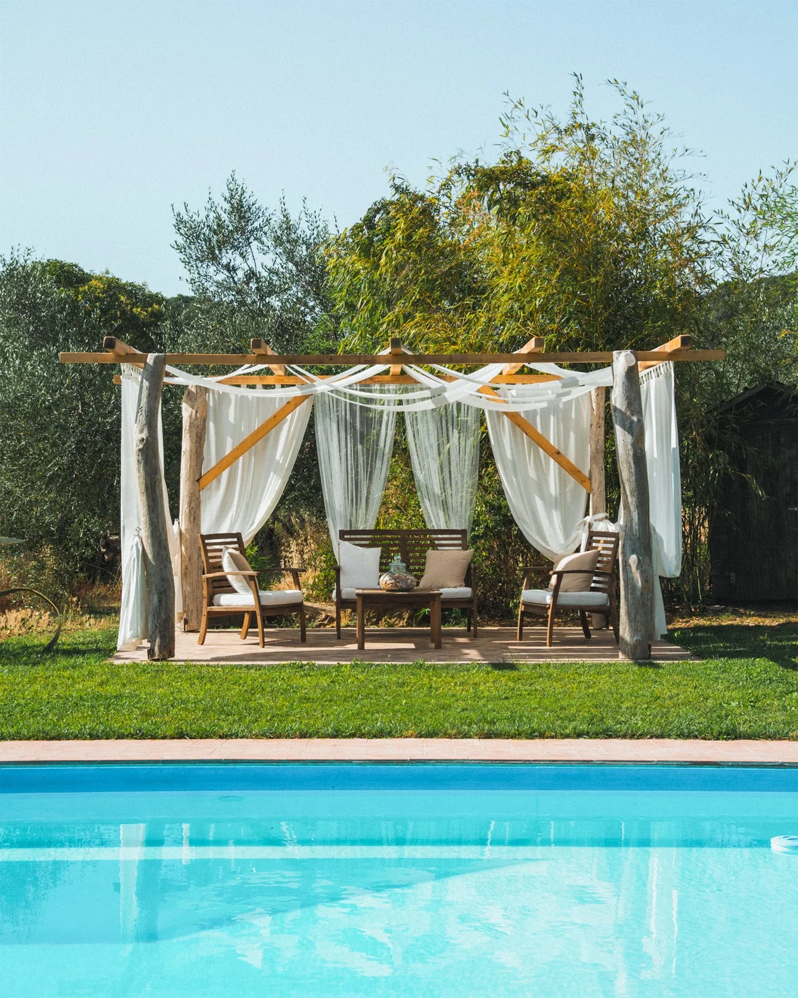 Gazebo con tende bianche e salotto da esterno sul prato, sul bordo della piscina azzurra: foto dallo shooting fotografico per la tenuta Colle Vento