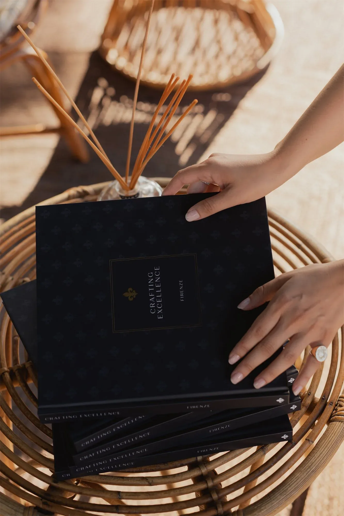 Mani che posano una pila di copie del libro Crafting Excellence. Firenze. su un tavolino di vimini: fotografia dell'evento di presentazione