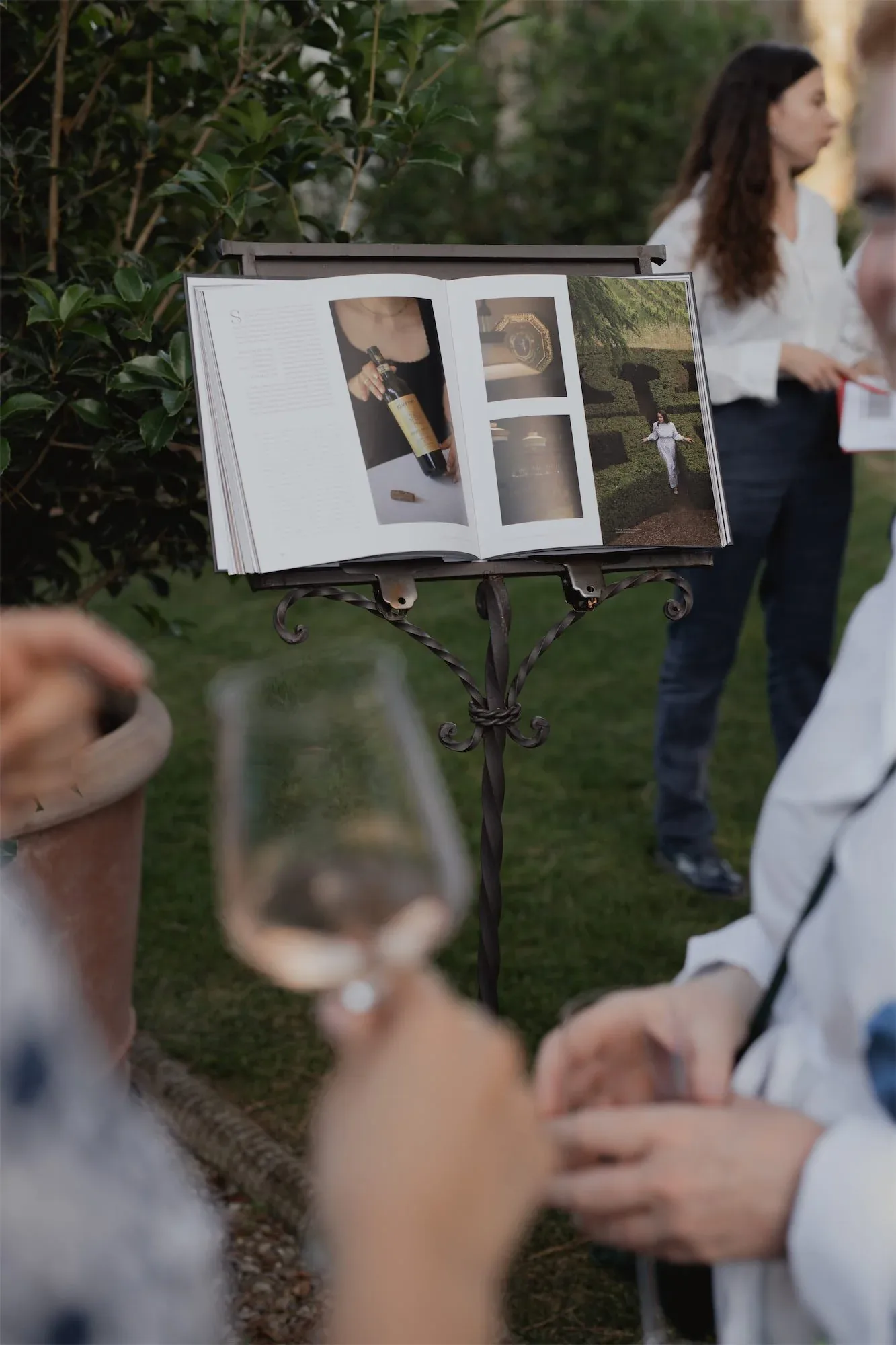 Libro aperto su un leggio in giardino, con gli ospiti intorno e un calice di vino: fotografia dell'evento di presentazione del libro