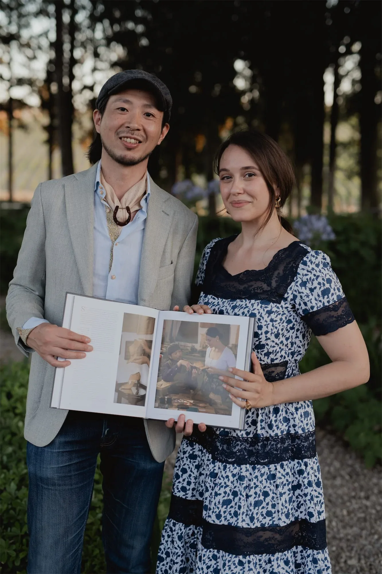 Un uomo con il cappello e una donna in abito a fiori posano con il libro aperto in giardino: fotografia dell'evento di presentazione