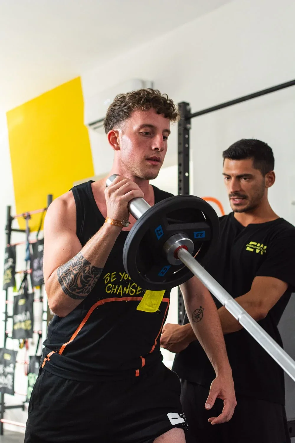 Ragazzo che solleva il bilanciere sulle spalle seguito da un istruttore con la maglia EW: foto dallo shooting in studio per Etruria Wellness