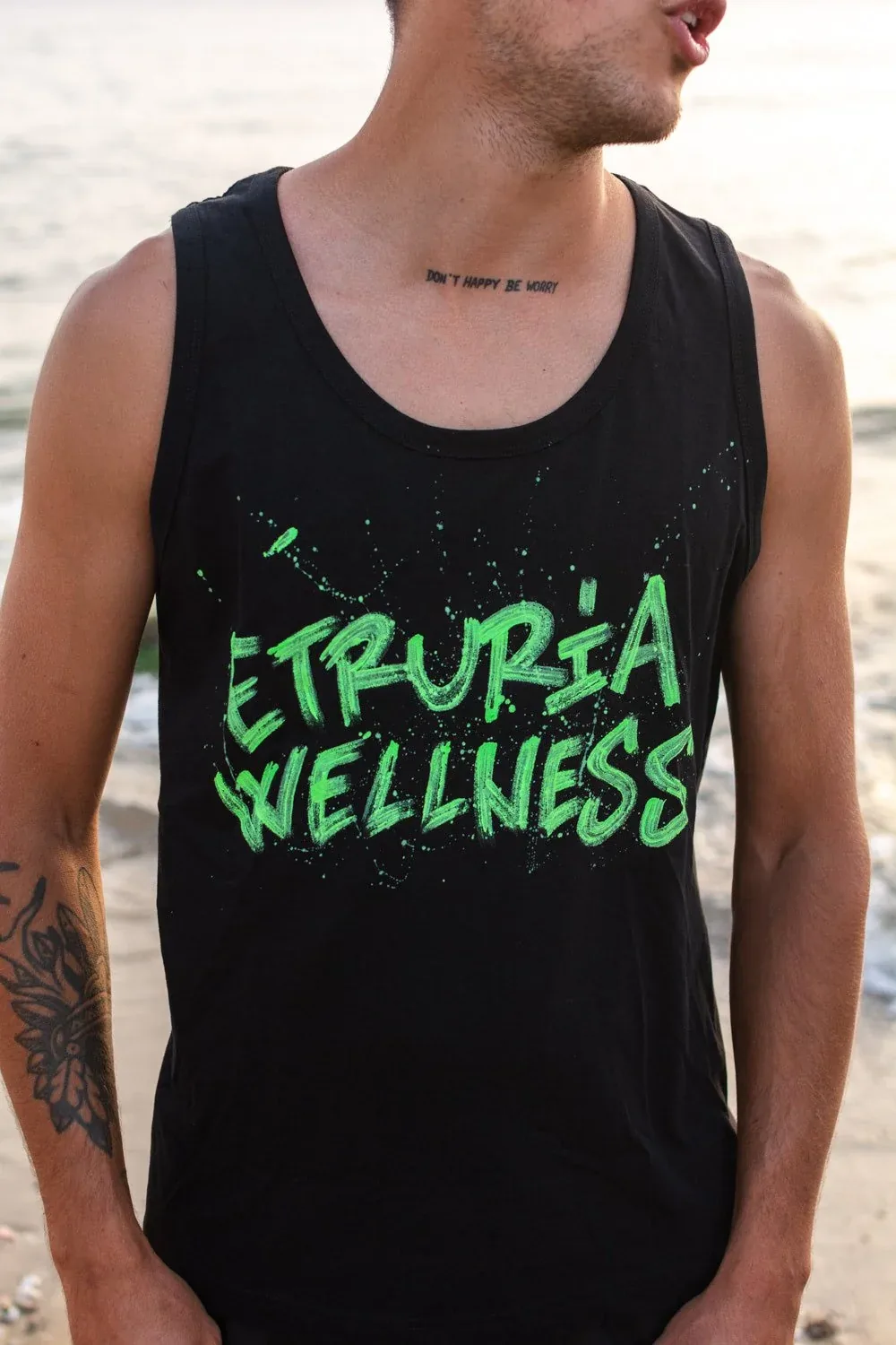 Canotta nera con la scritta Etruria Wellness dipinta in verde, indossata in spiaggia: concept di merchandising dallo shooting del brand