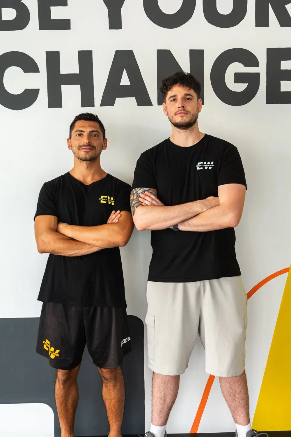 Due istruttori a braccia conserte con le maglie EW davanti al murale Be Your Change dipinto sulla parete: foto dallo shooting per Etruria Wellness