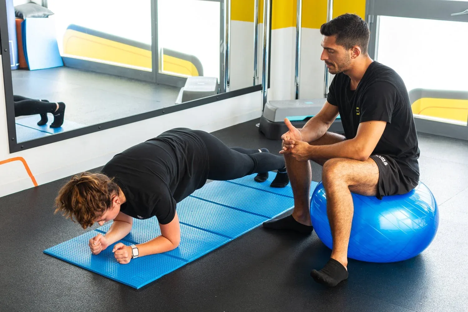 Ragazza in plank sul tappetino blu e istruttore seduto sulla fitball davanti allo specchio: foto dallo shooting in studio per Etruria Wellness