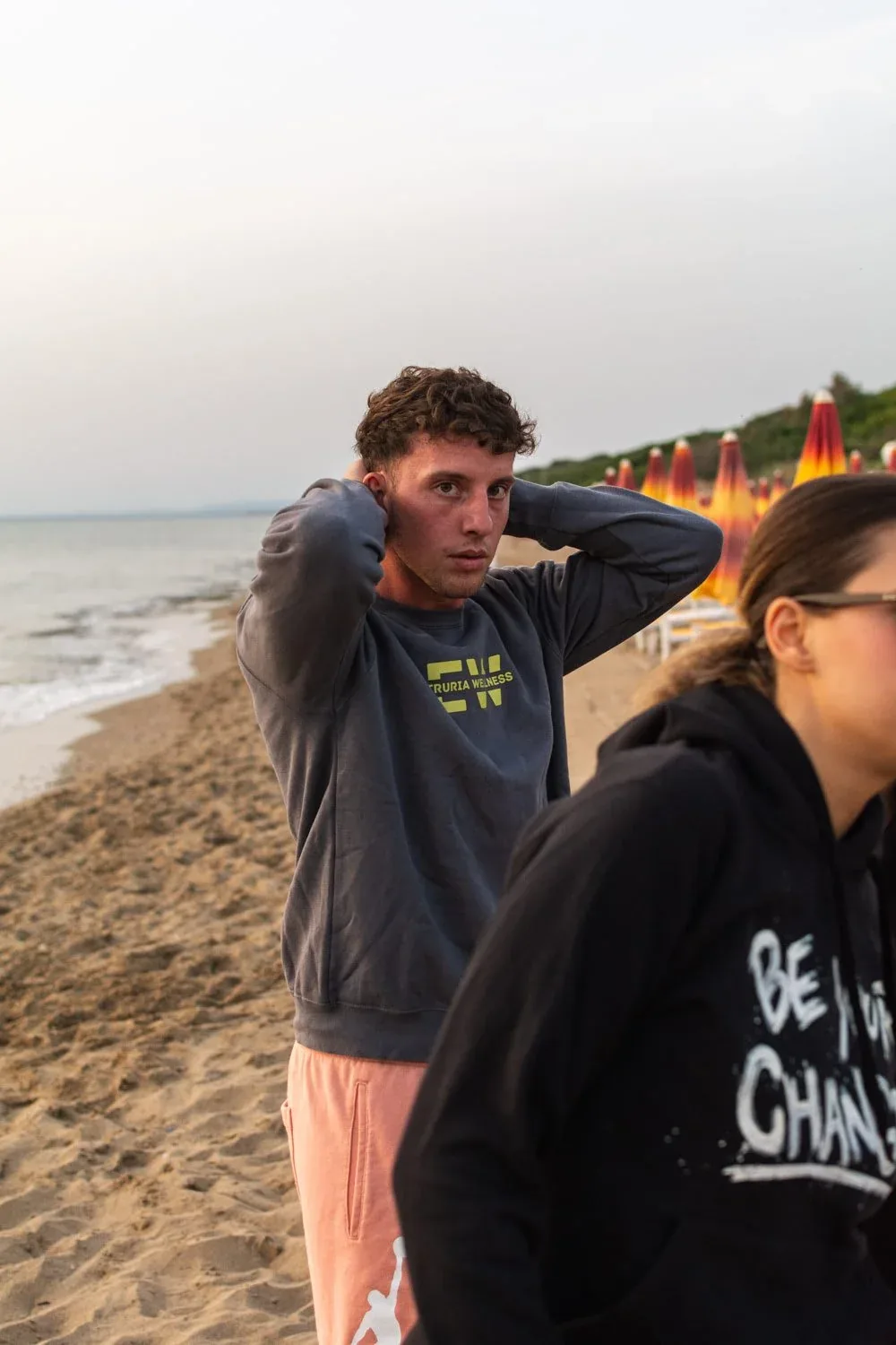 Ragazzo e ragazza con le felpe Etruria Wellness in spiaggia, sulla riva del mare con gli ombrelloni sullo sfondo: foto dallo shooting per Etruria Wellness