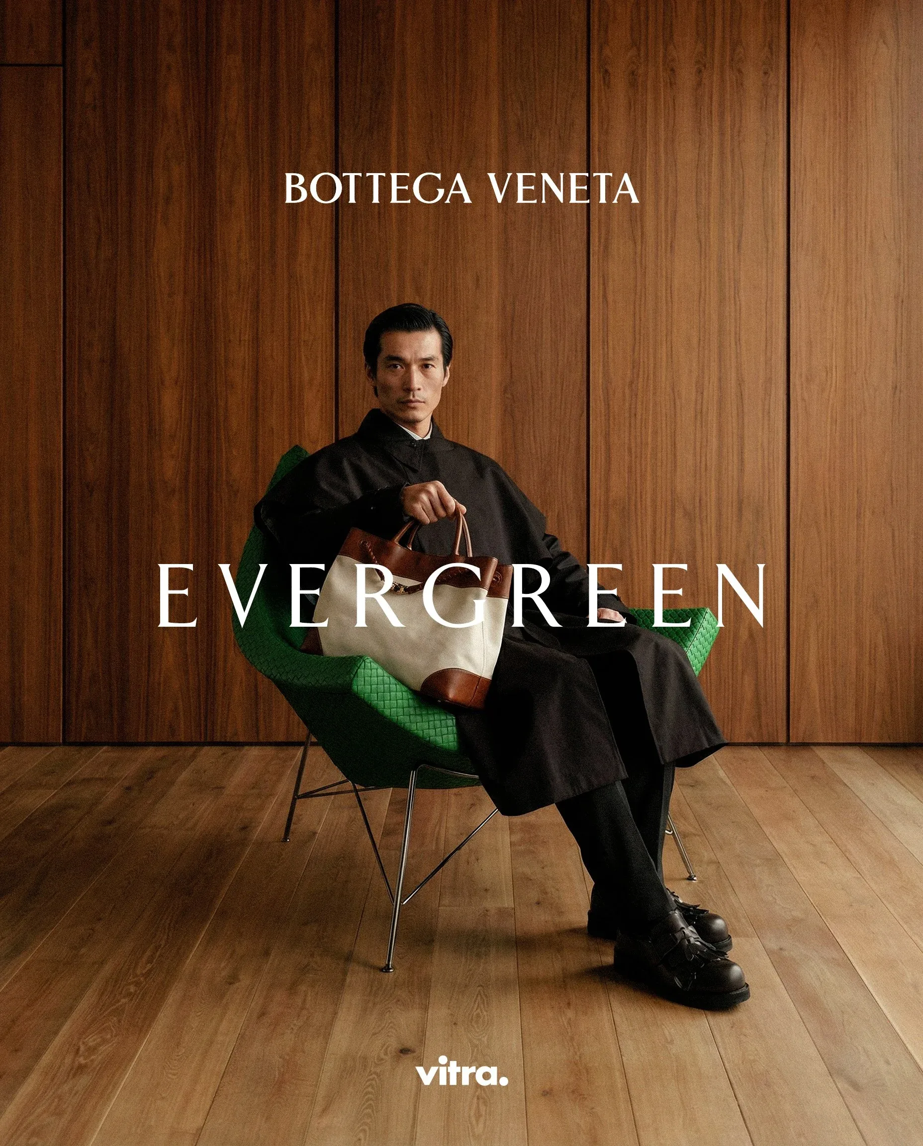 Uomo in cappotto scuro con una borsa seduto su una sedia verde davanti a una parete in legno, con le scritte Evergreen: visual della campagna moda