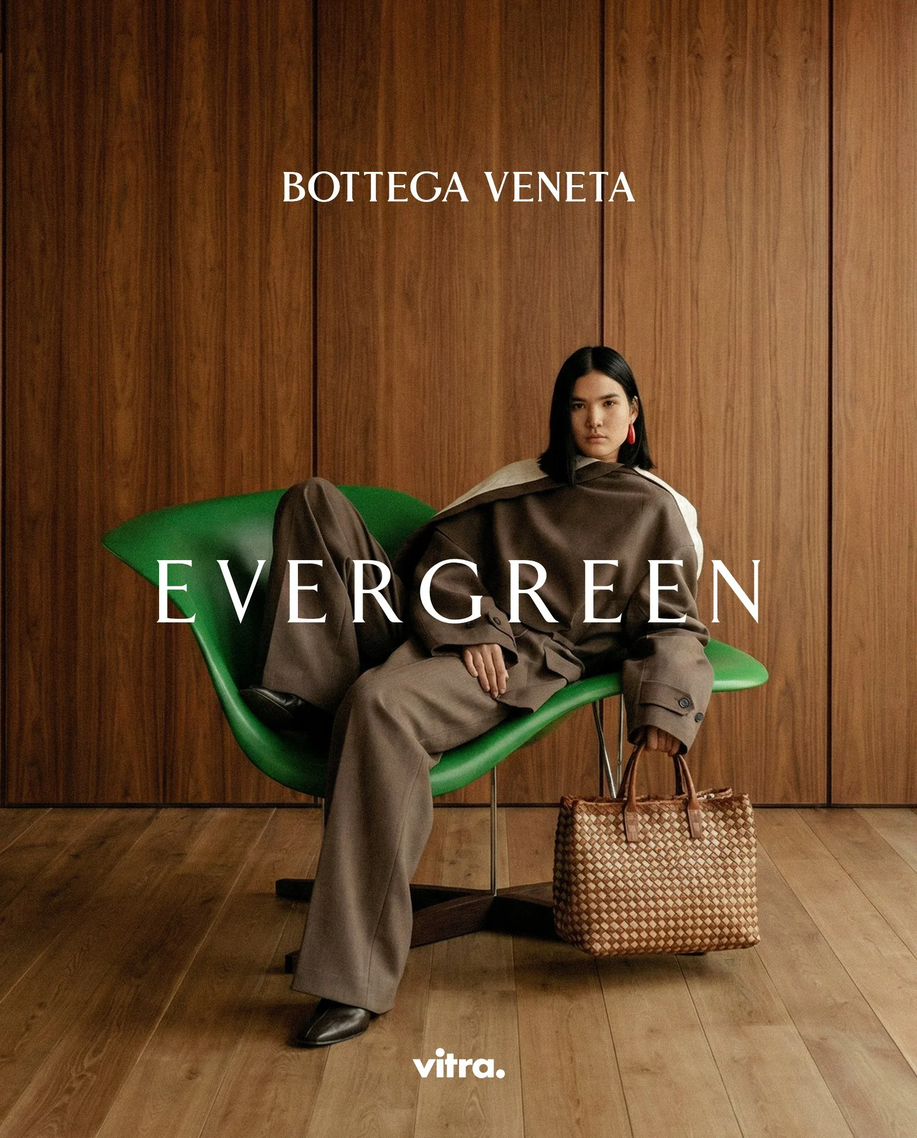 Donna sdraiata su una chaise longue verde con una borsa in intrecciato ai piedi, davanti a una parete in legno: visual della campagna moda Evergreen