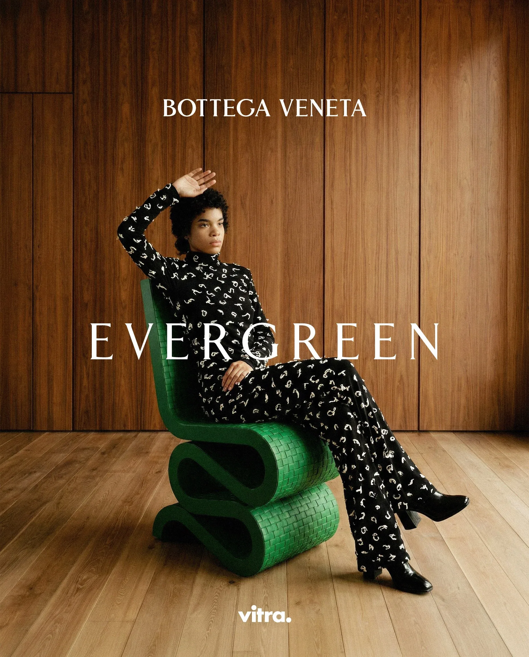 Donna in completo a fantasia bianca e nera seduta sulla sedia a onde verde, col braccio alzato, davanti a una parete in legno: visual della campagna moda