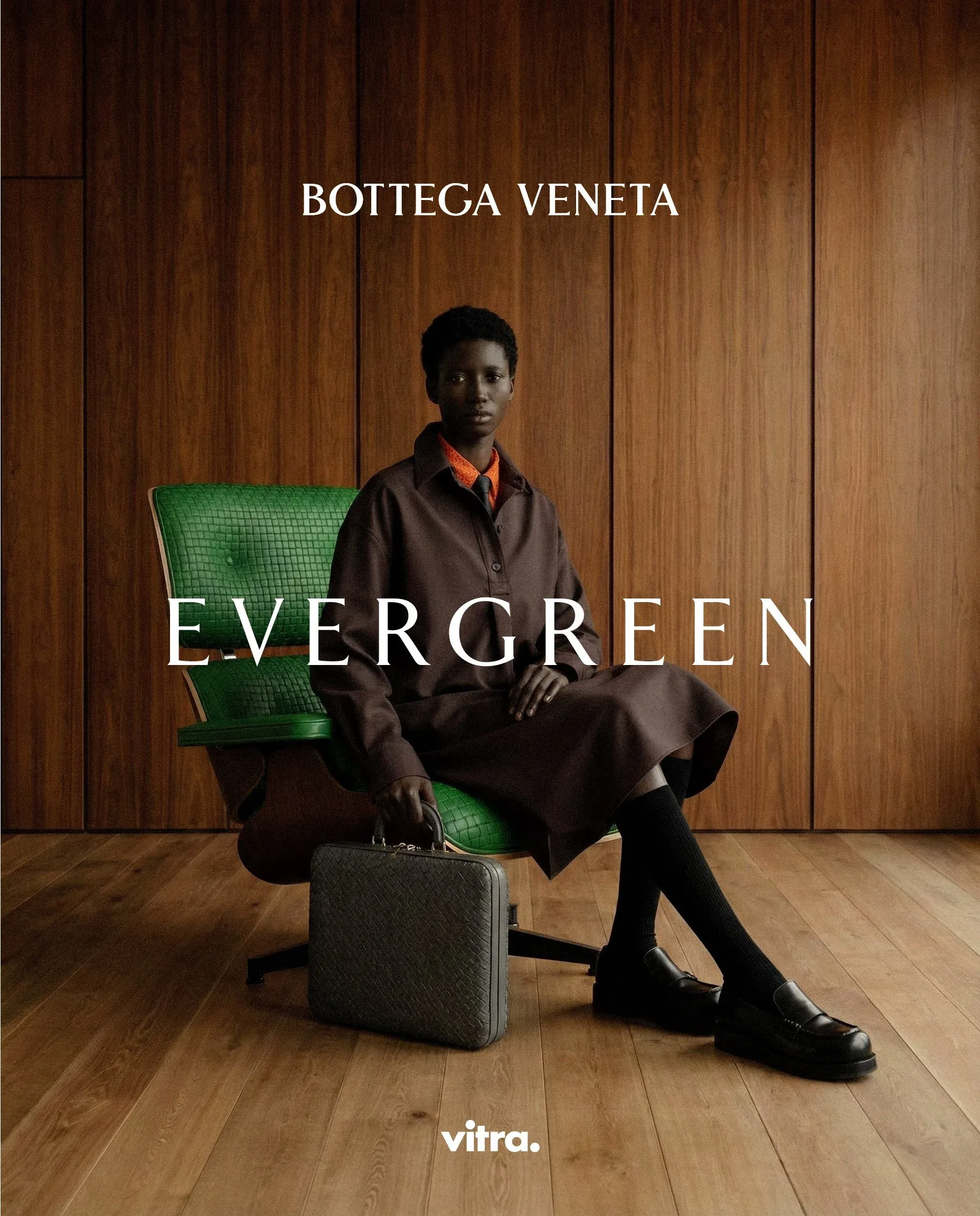 Persona in cappotto marrone seduta su una seduta verde con una valigetta in intrecciato accanto, davanti a una parete in legno: visual della campagna moda