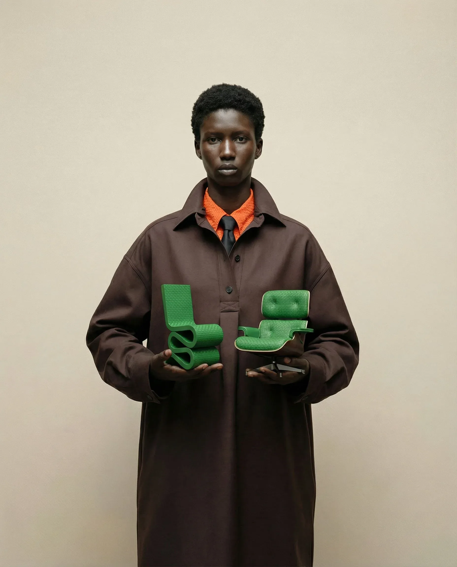 Persona in cappotto marrone e cravatta arancione che regge due miniature verdi di sedute Vitra: visual per The Evergreen Drop