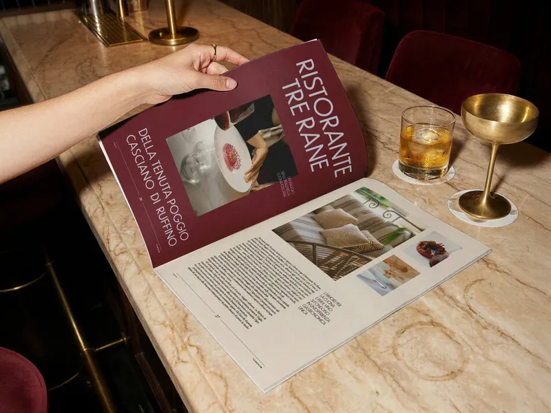 Tabloid F.U.C.K. Magazine aperto sull'articolo Ristorante Tre Rane, tenuto sopra un bancone in marmo con due cocktail: design editoriale