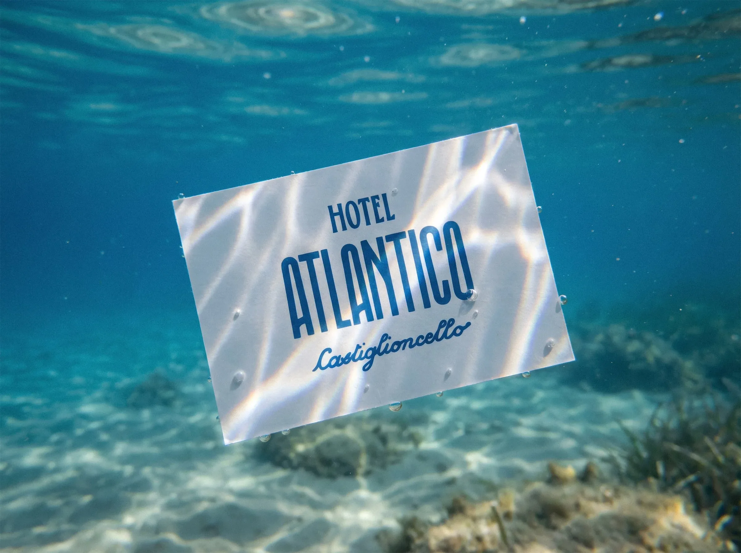 Biglietto da visita di Hotel Atlantico Castiglioncello con il logo blu, sospeso sott'acqua sopra un fondale di sabbia: mockup della visual identity