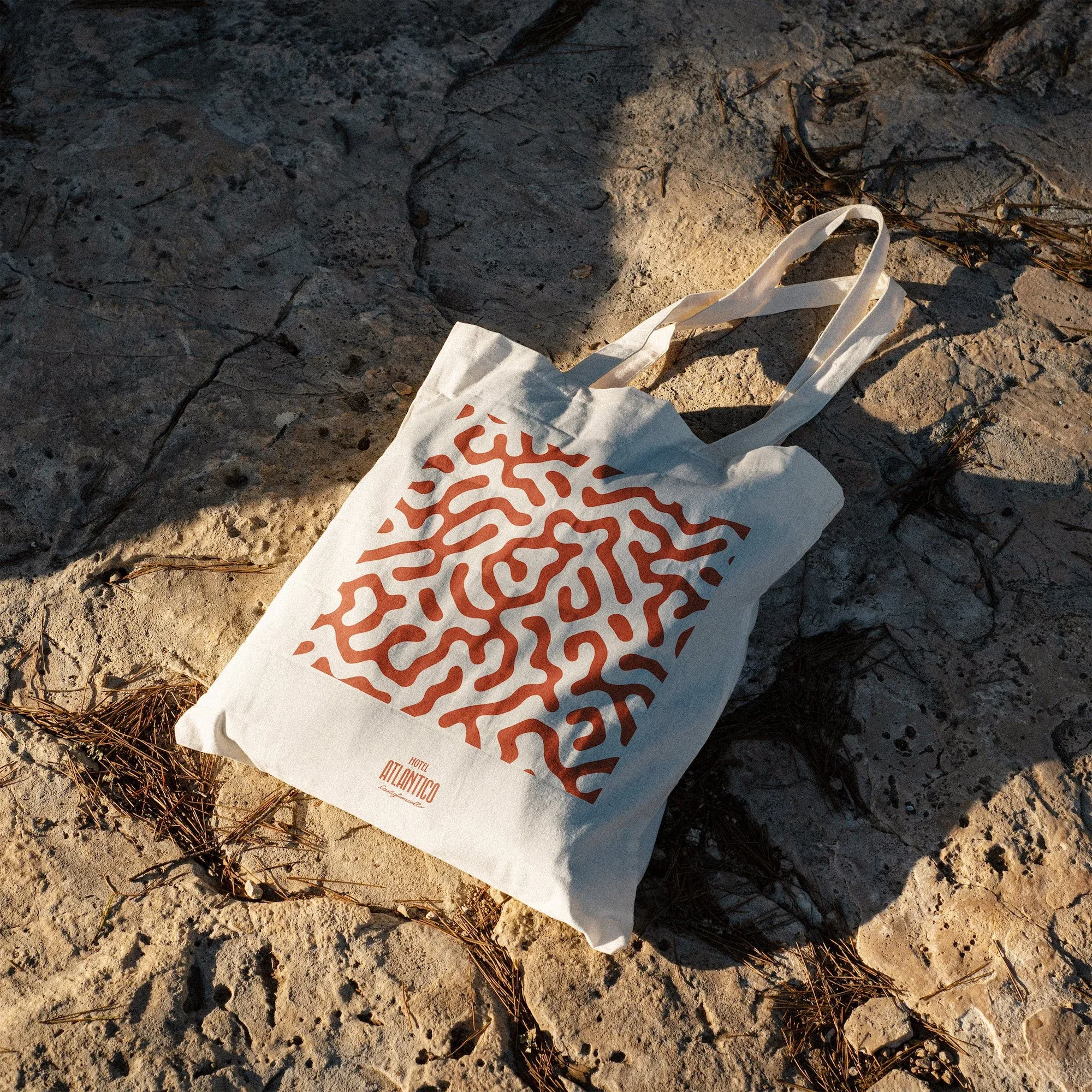 Tote bag in tela chiara con il pattern organico rosso di Hotel Atlantico, appoggiata su una roccia al sole: mockup di merchandising
