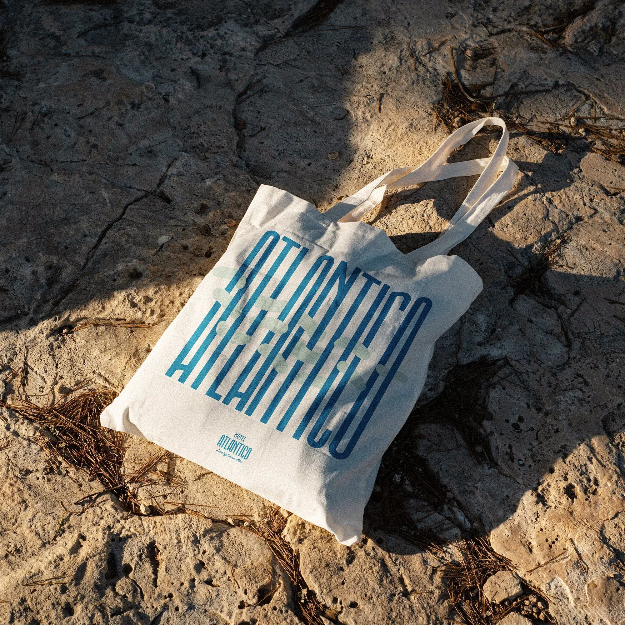 Tote bag in tela chiara con la scritta Atlantico in grandi lettere blu, appoggiata su una roccia al sole: mockup di merchandising per Hotel Atlantico