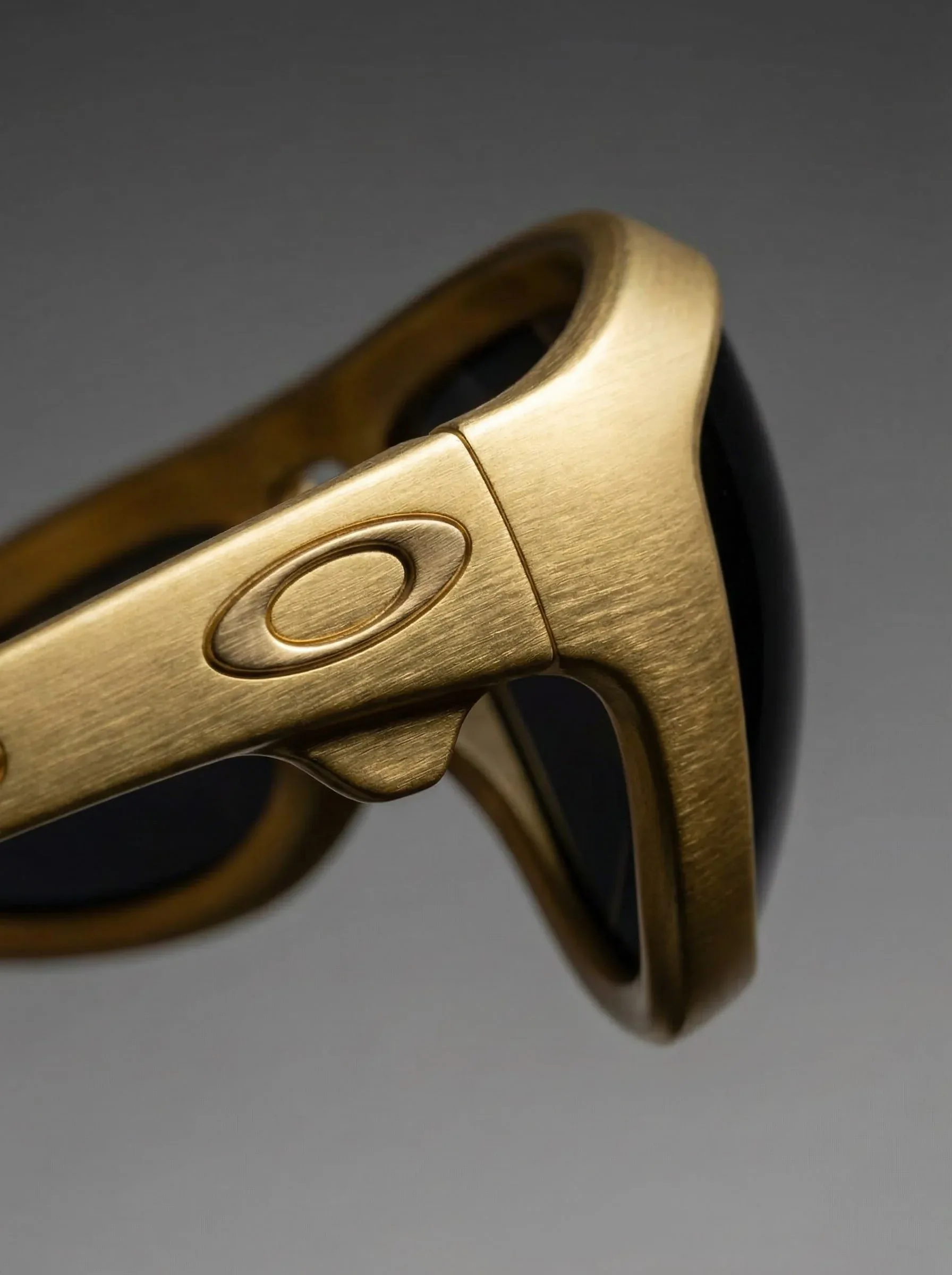 Dettaglio della stanghetta degli occhiali Oakley in oro spazzolato col logo in rilievo, su fondo grigio: design del prodotto