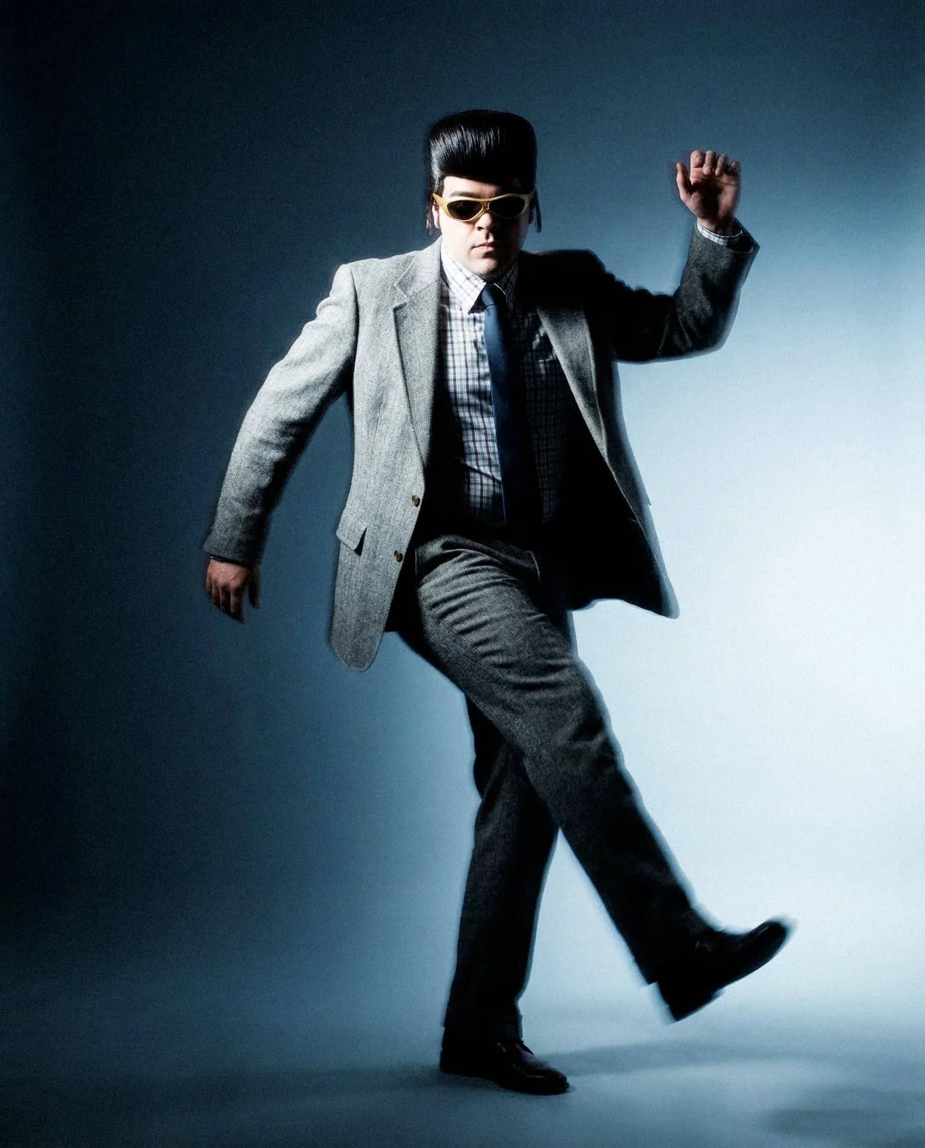 Tony Pitony a figura intera in giacca grigia e cravatta blu, colto in un passo di danza su fondo blu: shooting della campagna Oakley