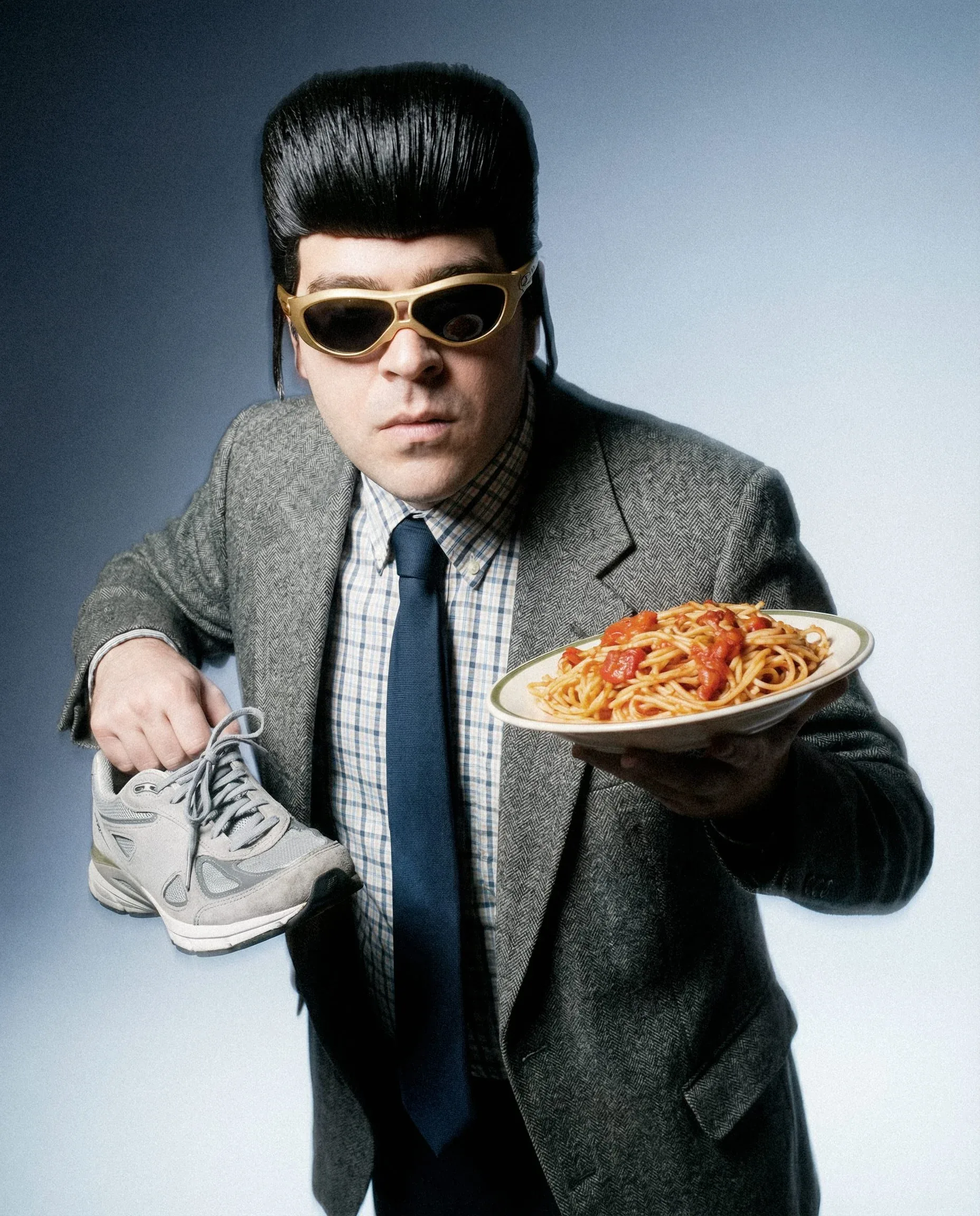 Tony Pitony con gli occhiali oro, una sneaker in una mano e un piatto di spaghetti nell'altra, su fondo azzurro: shooting della campagna Oakley