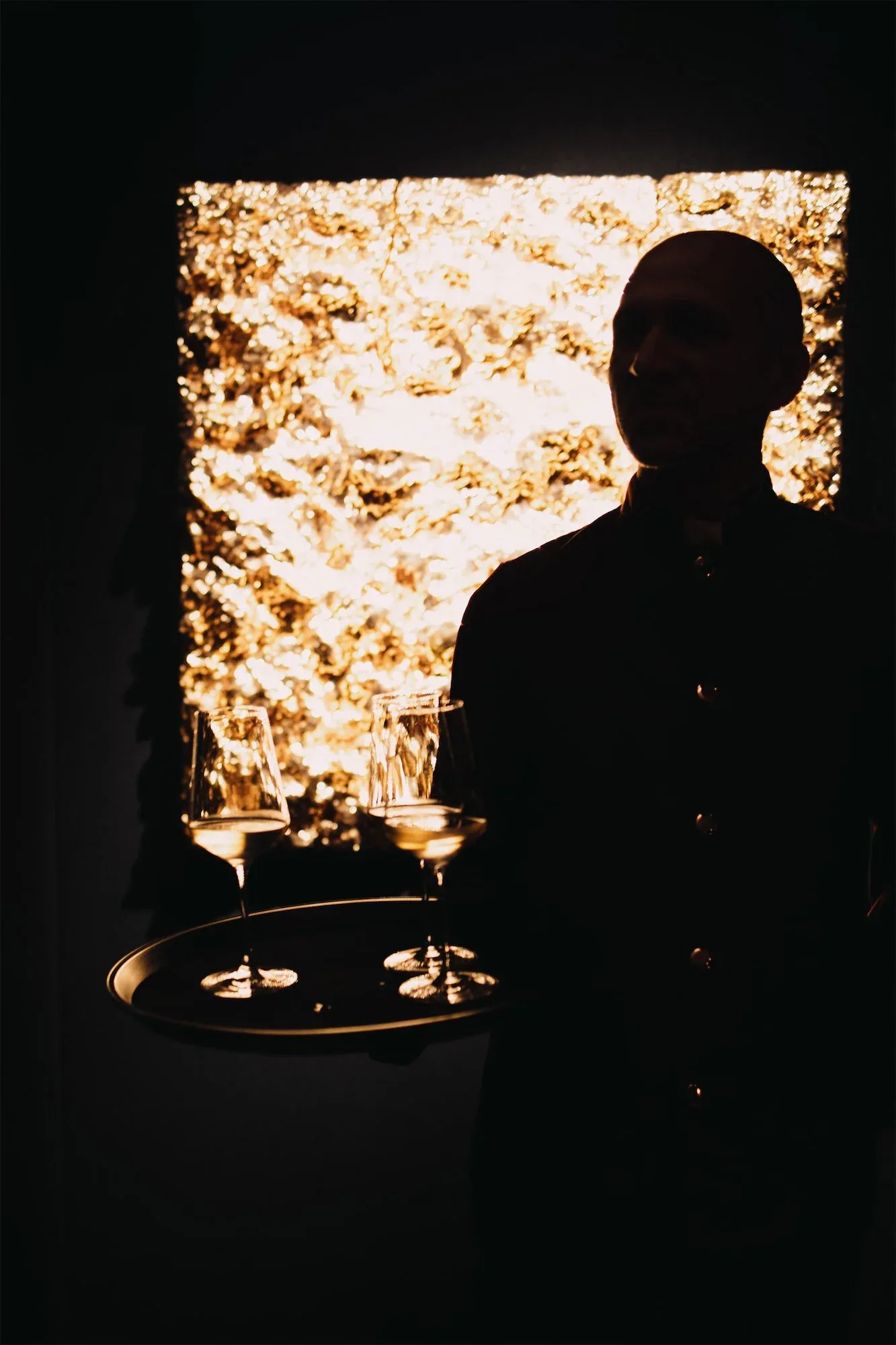 Un cameriere in controluce porta un vassoio di calici davanti a un pannello d'oro illuminato: fotografia dell'evento ORObyRUFFINO 2024