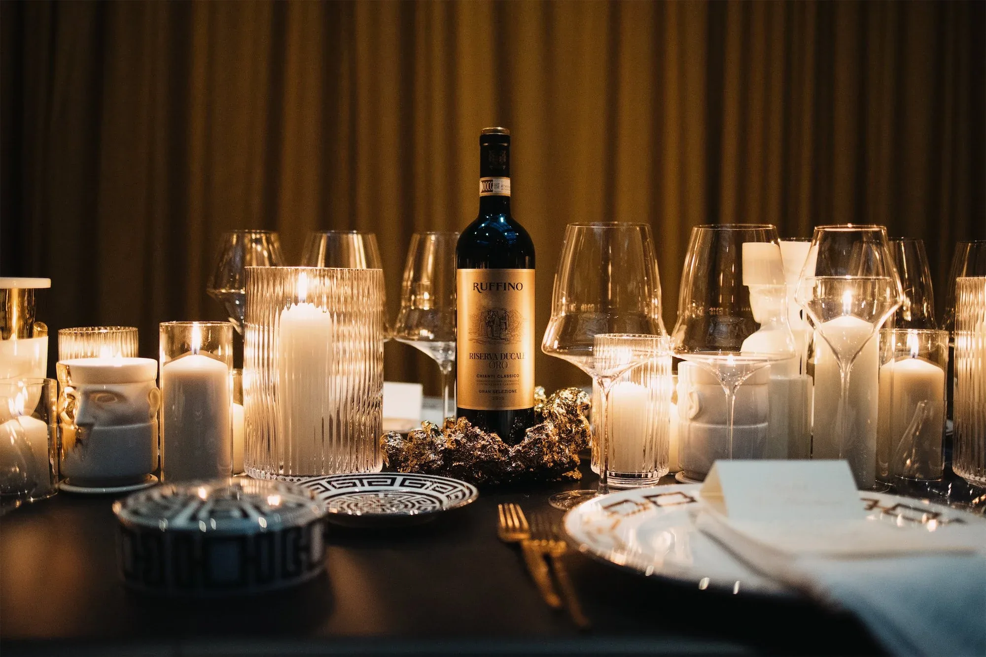 Tavola apparecchiata con candele accese, calici e una bottiglia Ruffino davanti a tende dorate: fotografia della cena di ORObyRUFFINO 2024